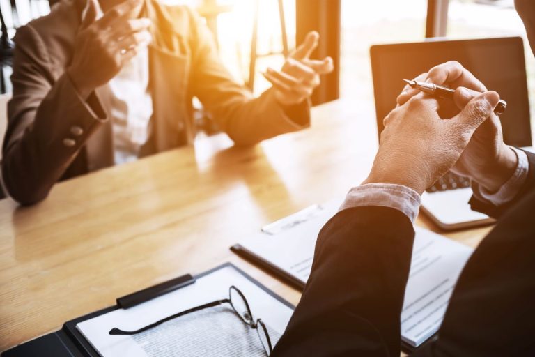 Two professionals discussing documents across a desk with a laptop and clipboard during a business meeting.