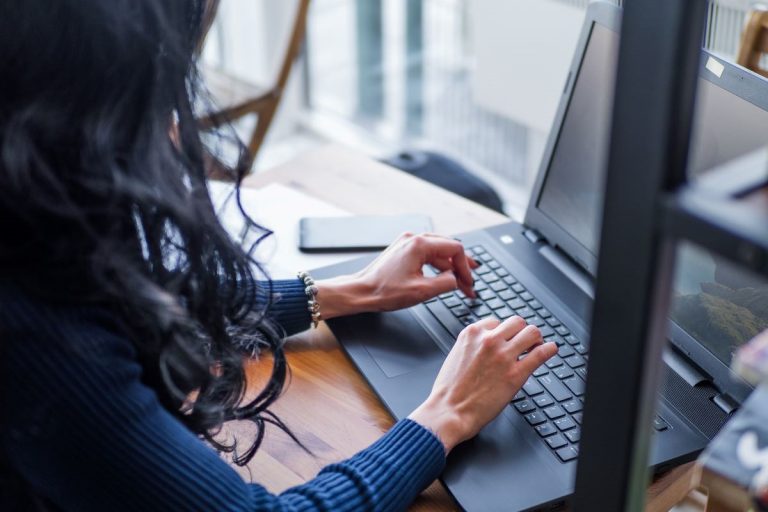 Person typing on a laptop at a desk near a window with a smartphone beside the computer.