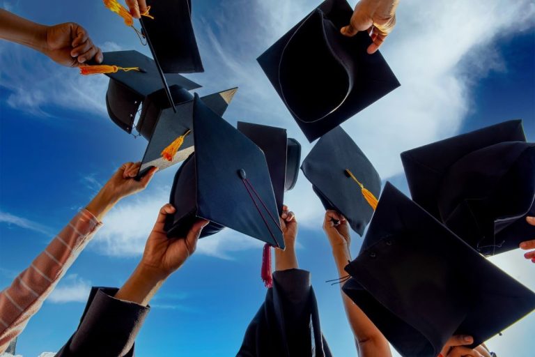 Graduates raising black caps with tassels into the air against a bright blue sky during a graduation celebration.