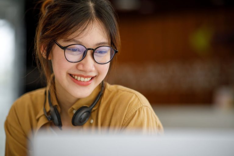Smiling woman with glasses wearing headphones around her neck while looking at a laptop screen.