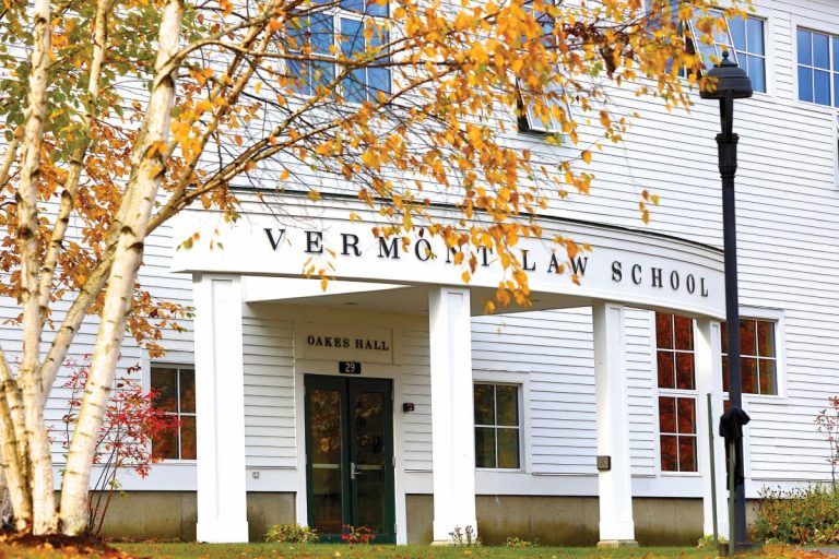 Entrance of Vermont Law School with white columns and a sign reading "Vermont Law School" above the doorway, surrounded by autumn trees with orange and yellow leaves. The scene highlights a quiet campus setting during fall.