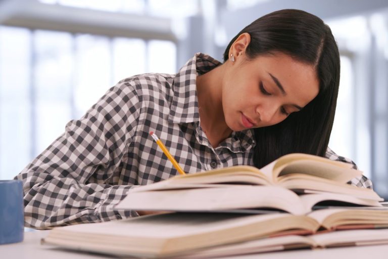 Student writing in a notebook while studying with open textbooks at a desk in a bright study space.