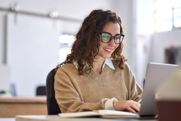 Woman with curly hair and glasses smiling while typing on a laptop at a desk in a bright office.