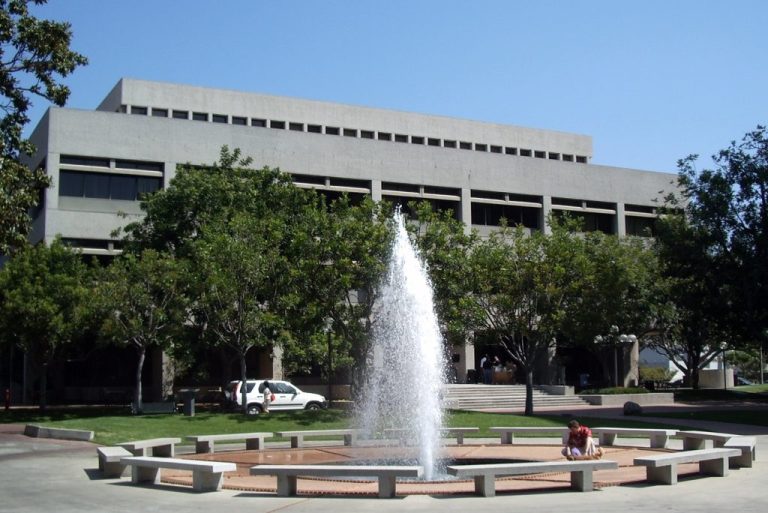 USC Gould School of Law building with a large fountain spraying water in a circular courtyard surrounded by trees and benches.