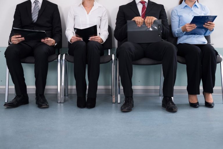 Four job candidates in business attire sitting in chairs holding folders and clipboards while waiting for an interview.