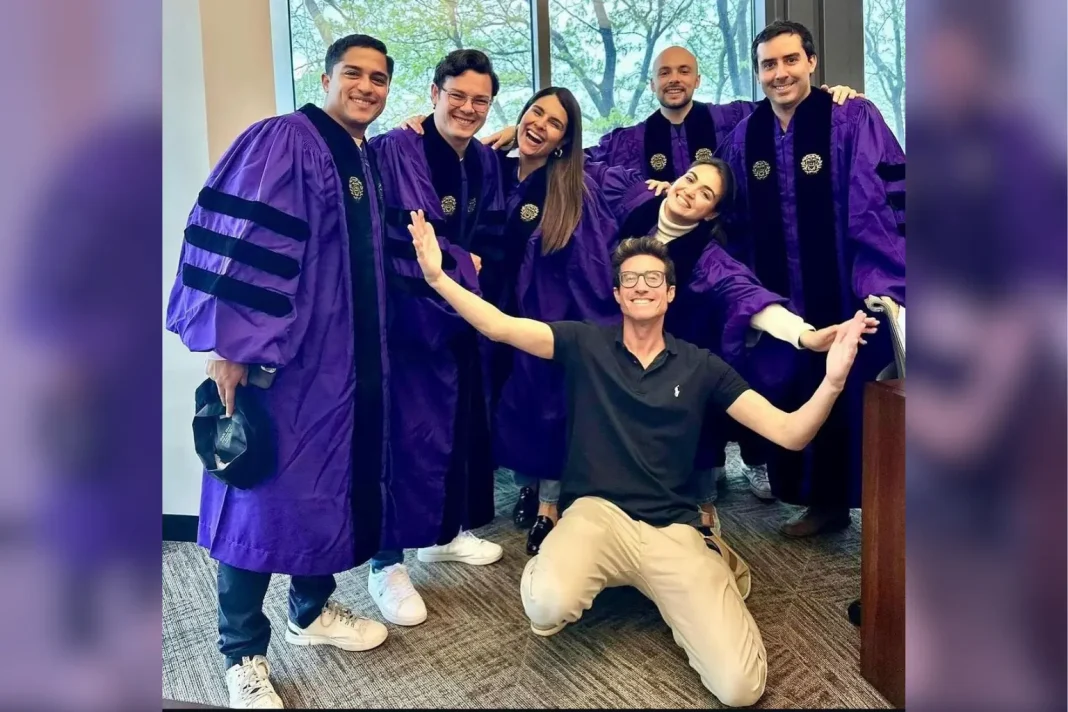 Group of smiling law graduates in purple academic robes pose together indoors with Joshua Alter kneeling in front with his arms raised. Large windows behind them show trees outside as they celebrate the moment together.