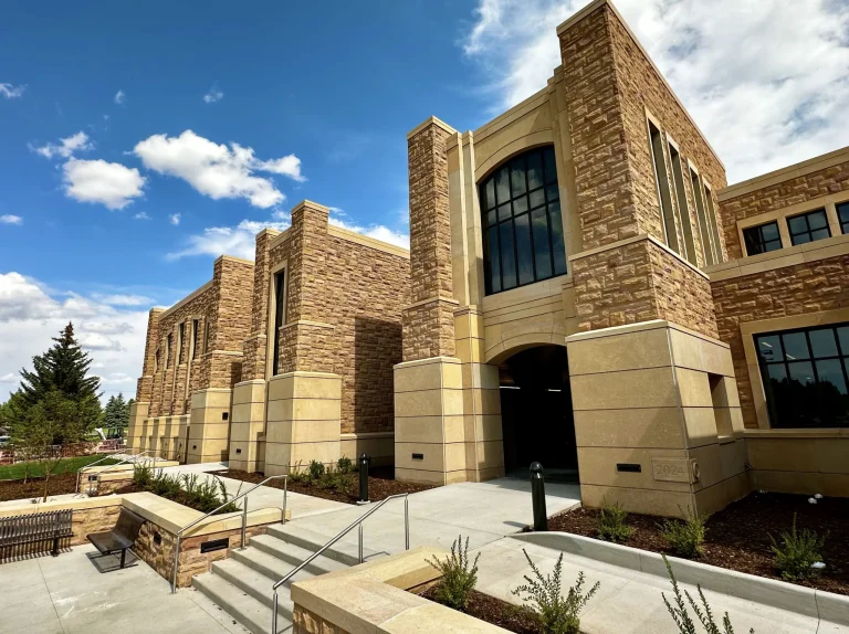 Stone exterior of the University of Wyoming College of Law building with tall vertical windows and a large arched entrance. Concrete steps and landscaped walkways lead up to the new building marked “2024” under a bright blue sky with scattered clouds.