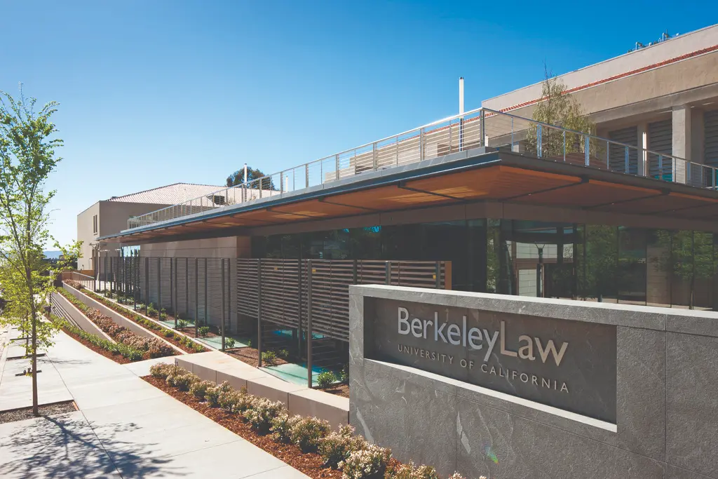 A modern building with clean lines and large glass windows, identified by a sign reading “Berkeley Law, University of California.” The landscaped walkway and contemporary architecture suggest a prestigious academic law school campus.