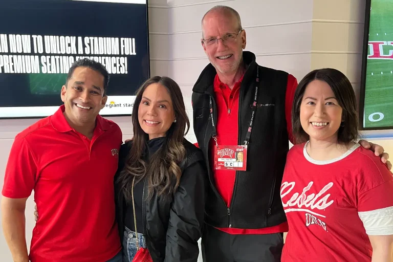 Four people wearing UNLV apparel standing together and smiling indoors, with stadium seating information displayed on screens in the background.