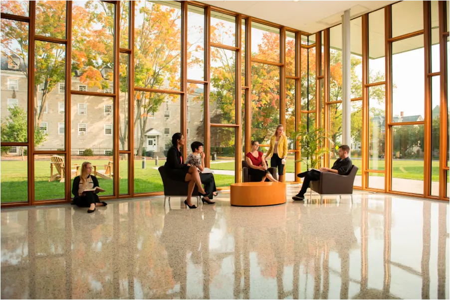 A group of students and professionals sitting and conversing in a bright, modern lounge with floor-to-ceiling windows, overlooking a campus with autumn-colored trees.