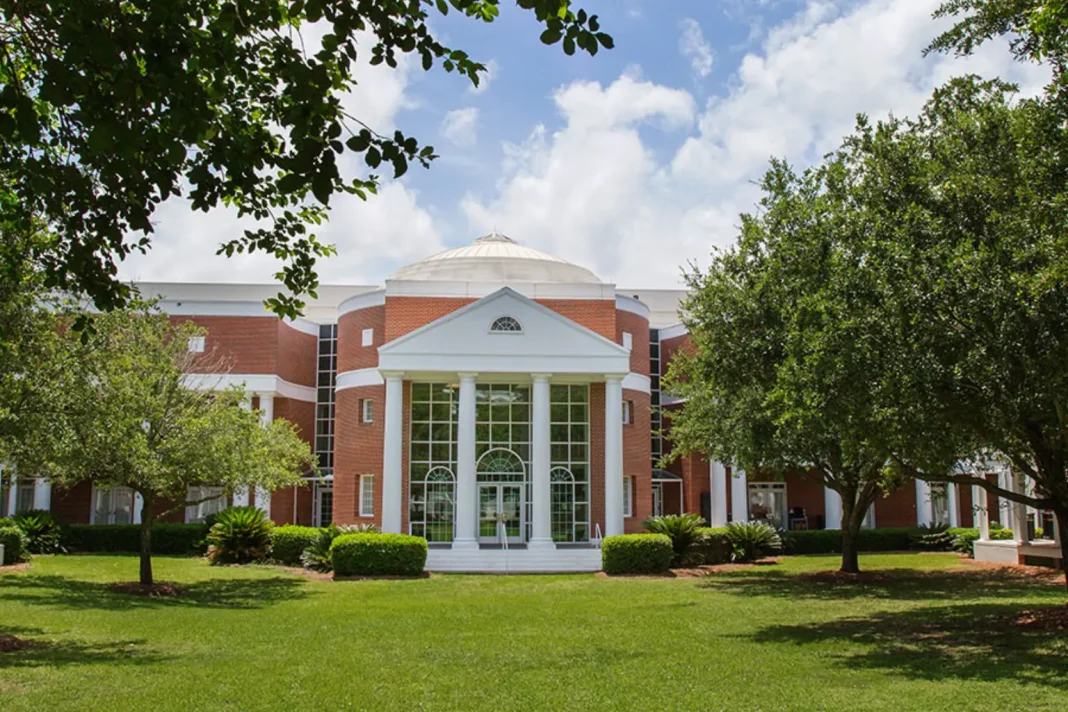 Red brick academic building with white columns and a domed roof surrounded by trees and a green lawn on the Florida State University campus. The symmetrical entrance features tall windows and a classical portico under a bright blue sky with scattered clouds.