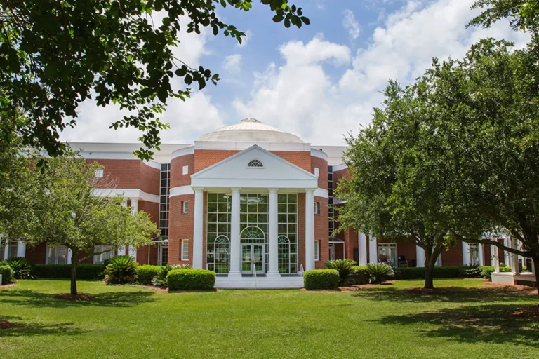 Red brick academic building with white columns and a domed roof surrounded by trees and a green lawn on the Florida State University campus. The symmetrical entrance features tall windows and a classical portico under a bright blue sky with scattered clouds.