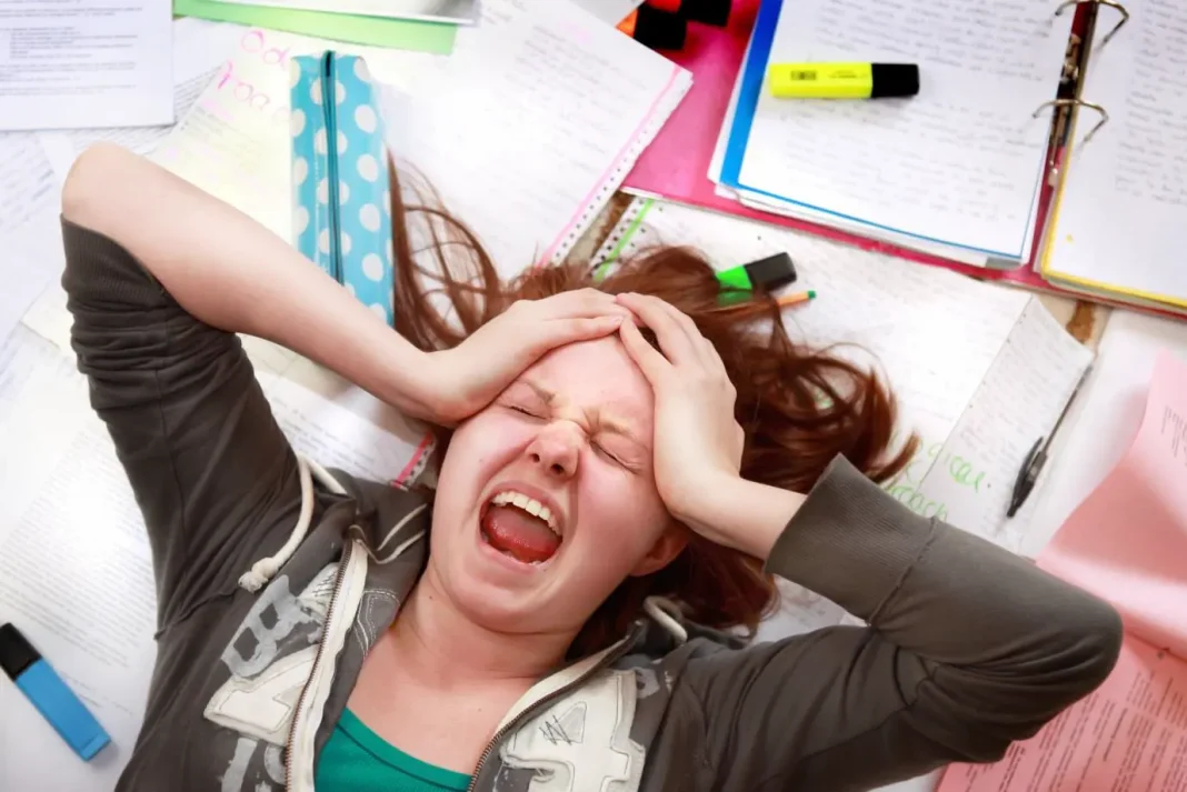 Overwhelmed student lying on a desk surrounded by scattered papers, notebooks, and highlighters, holding their head and screaming in frustration from studying stress.