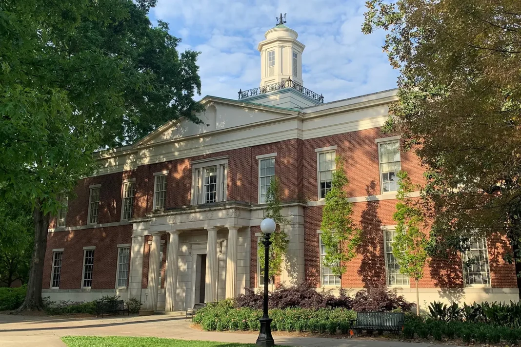The University of Georgia School of Law building, a red-brick academic hall with white columns and a central cupola, surrounded by trees and greenery on the UGA campus.