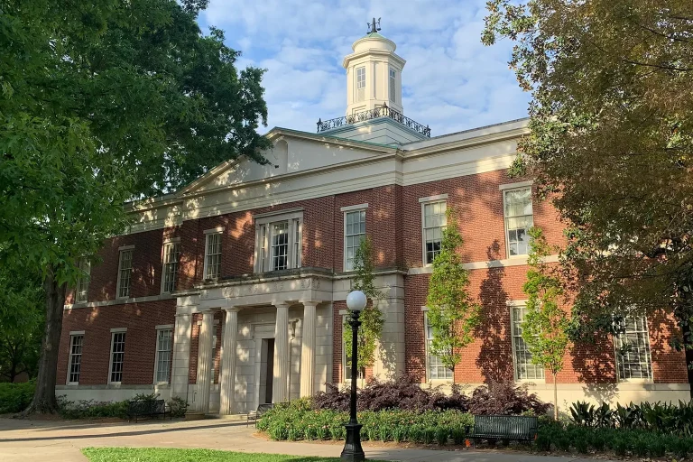 The University of Georgia School of Law building, a red-brick academic hall with white columns and a central cupola, surrounded by trees and greenery on the UGA campus.