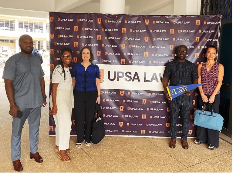 Five people stand in front of a backdrop repeating the text “UPSA LAW” and the UPSA crest during a campus visit or partnership event. One person holds a blue pennant that reads “UC Law San Francisco.”