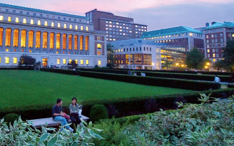 Columbia University law school buildings illuminated at dusk overlooking a large campus lawn with students sitting and walking nearby.