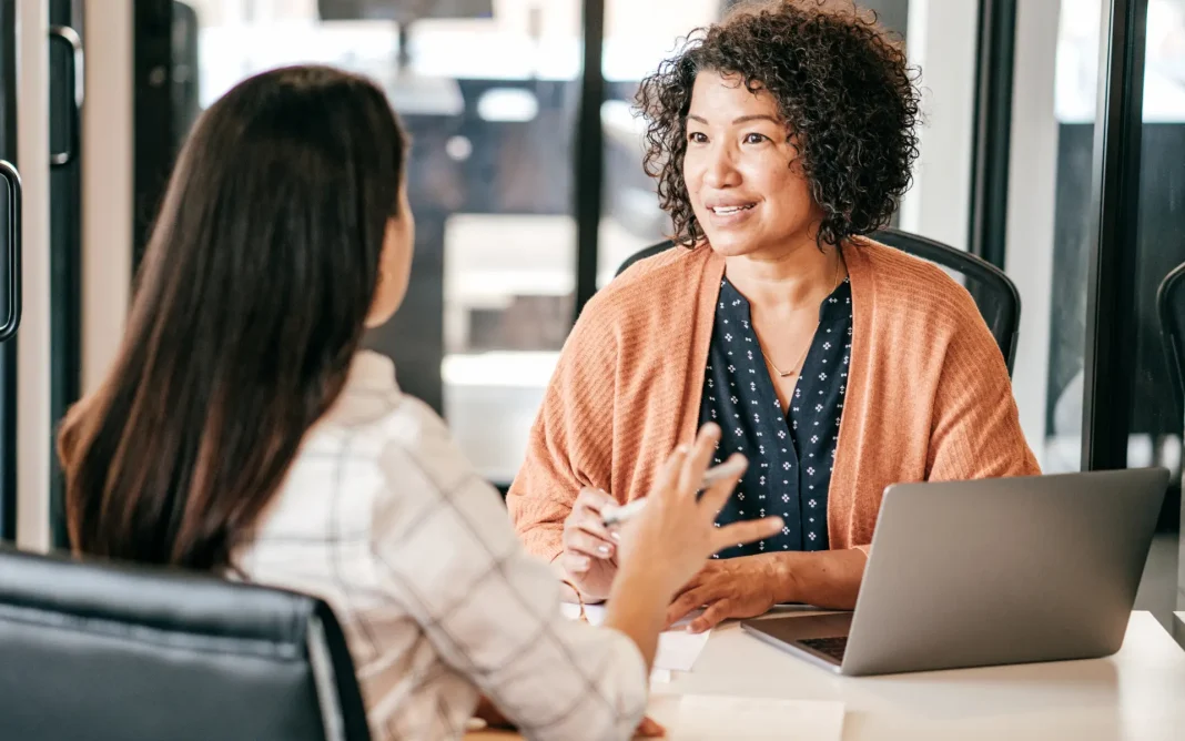 Two women sitting at a desk having a conversation in an office, with one taking notes beside a laptop.