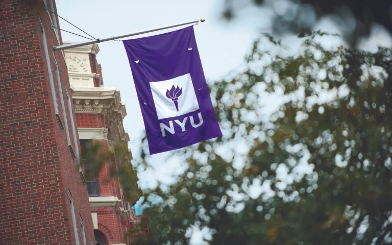 Purple NYU banner with torch logo hanging from a building, with trees and part of a campus building in the background.
