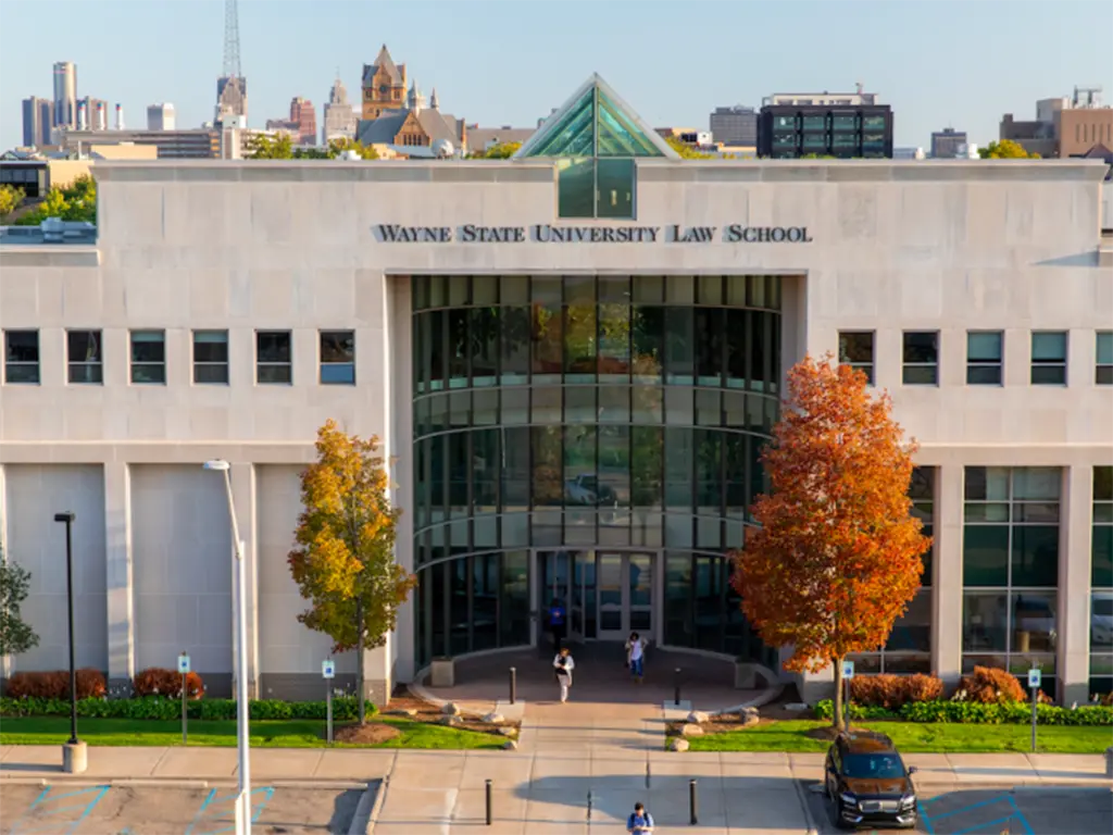 Front entrance of a modern academic building labeled &ldquo;Wayne State University Law School&rdquo; with a tall glass atrium and triangular skylight above the doorway. Two people walk near the entrance while autumn trees with orange leaves frame the building against a city skyline in the background.