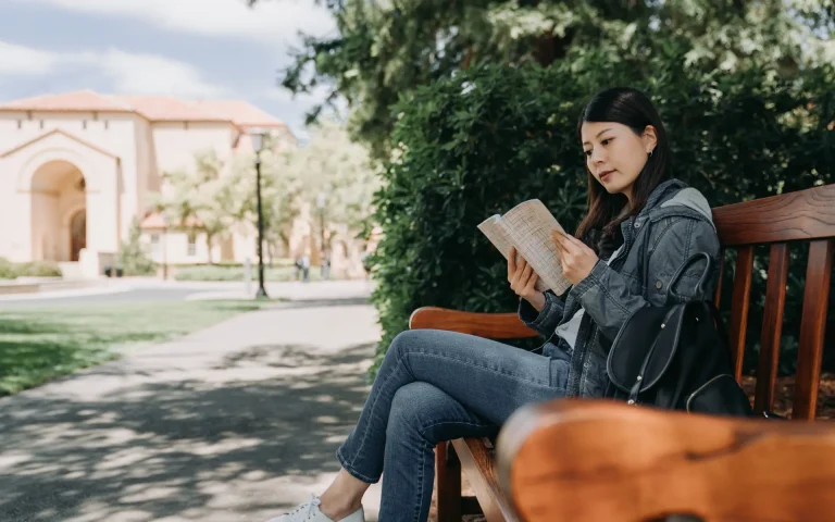 Student sitting on a wooden bench on a university campus reading a small notebook, with trees and a campus building in the background.