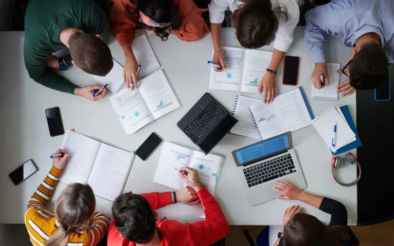 Overhead view of a group of students gathered around a table studying together with laptops, notebooks, phones, and printed charts while taking notes and discussing materials.