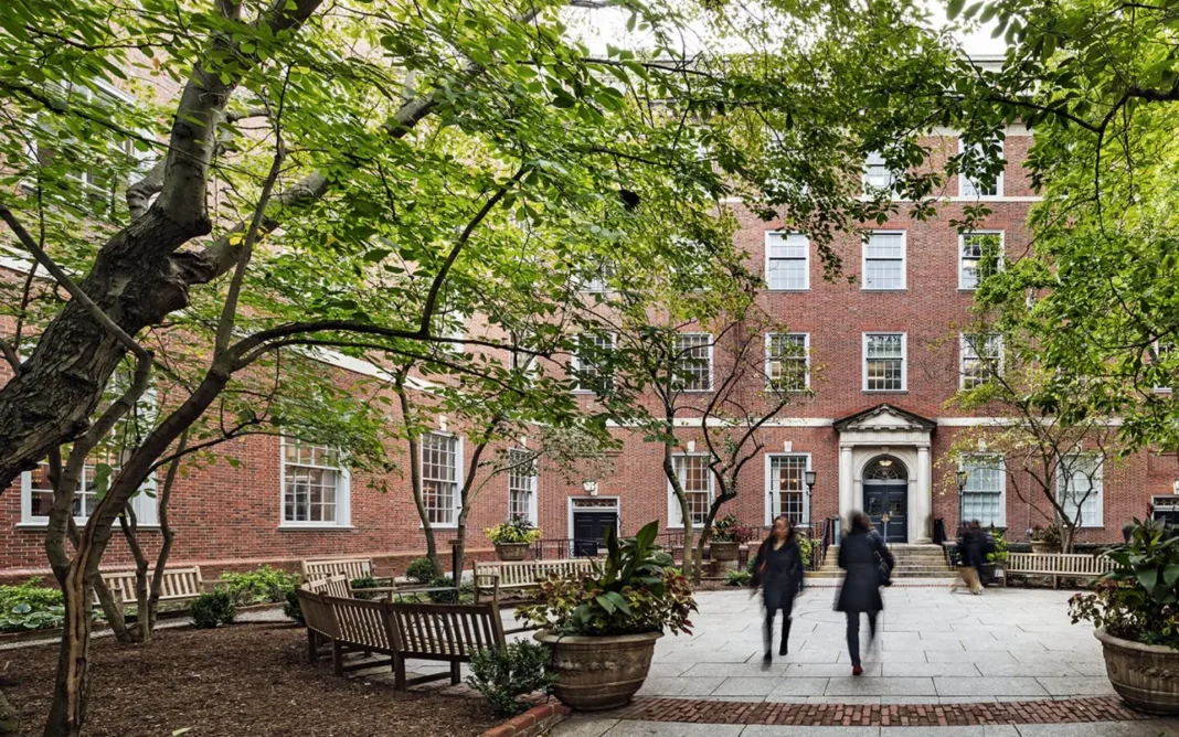 Tree-lined courtyard with benches and planters in front of a red-brick academic building, with a few blurred pedestrians walking across the paved path.