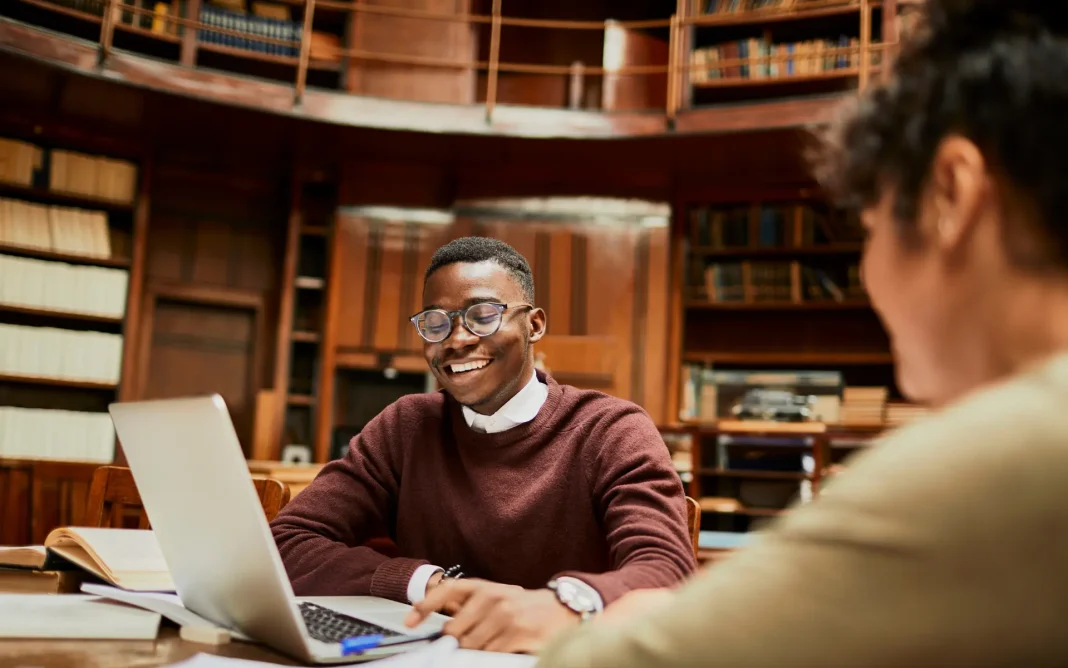Two students collaborating at a table in a library, one using a laptop while bookshelves and study materials surround them.