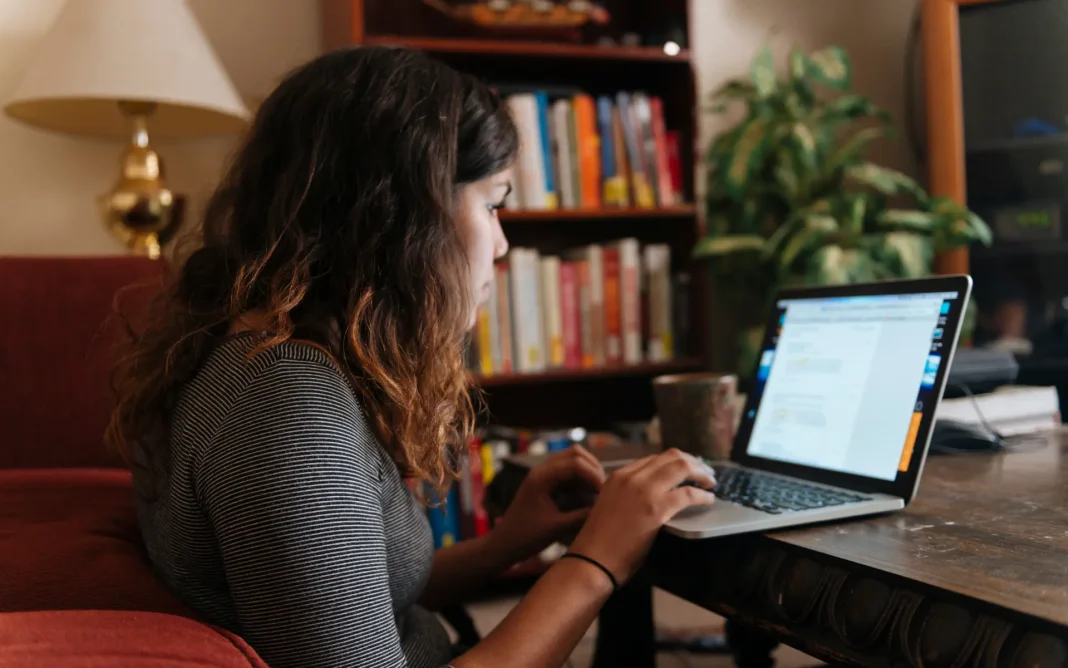 Student working on a laptop at a desk in a cozy home study space with bookshelves and a lamp in the background.