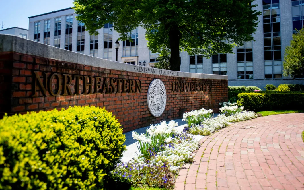 Brick wall sign reading “Northeastern University” at Krentzman Quadrangle with campus buildings, trees, and landscaped flowers in the background.