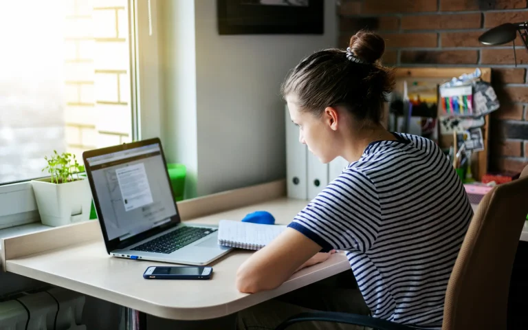 Student sitting at a desk by a window studying on a laptop with a notebook and phone nearby.