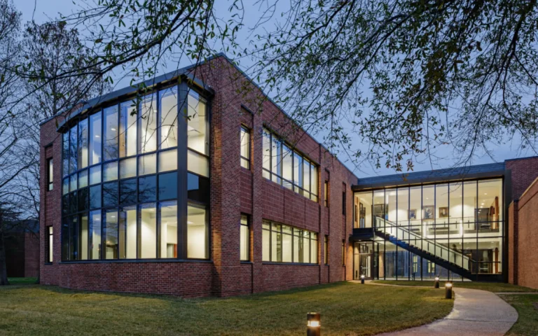 Modern red-brick law school building with large glass windows and a glass stairway entrance, illuminated at dusk and surrounded by trees and a curved walkway.