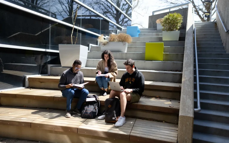 Three students sitting on outdoor campus steps studying together, reading and using a laptop with backpacks beside them.