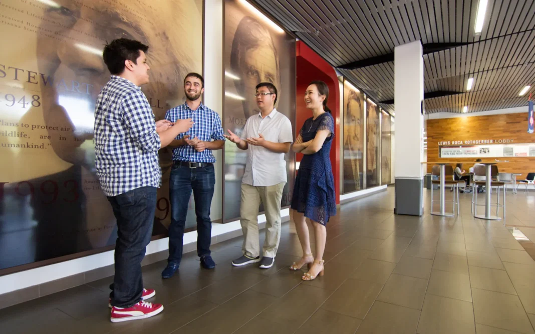 Four students standing and talking together in a modern university lobby hallway with large portrait displays on the walls.