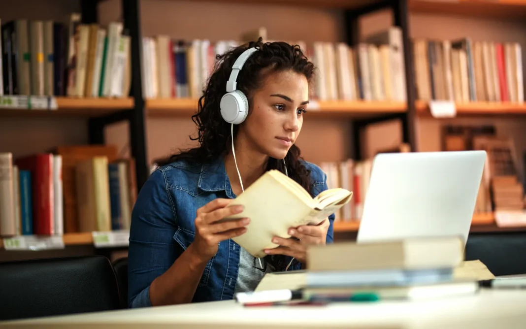 Student wearing headphones studying in a library, holding an open book while using a laptop at a table with bookshelves in the background.