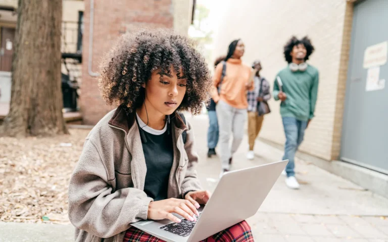 College student with curly hair sitting outdoors using a laptop while other students walk by in the background on a campus pathway.