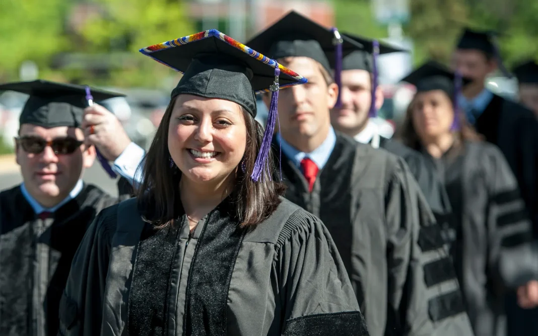 Group of graduates in caps and gowns standing in line outdoors during a graduation ceremony, with a smiling graduate wearing a decorated cap in the foreground.