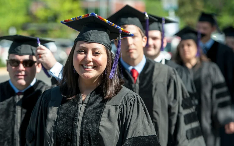 Group of graduates in caps and gowns standing in line outdoors during a graduation ceremony, with a smiling graduate wearing a decorated cap in the foreground.
