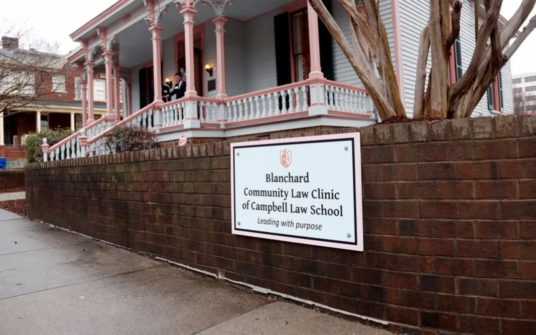 A sign reading “Blanchard Community Law Clinic of Campbell Law School” mounted on a brick wall in front of a historic house-style building with a porch.