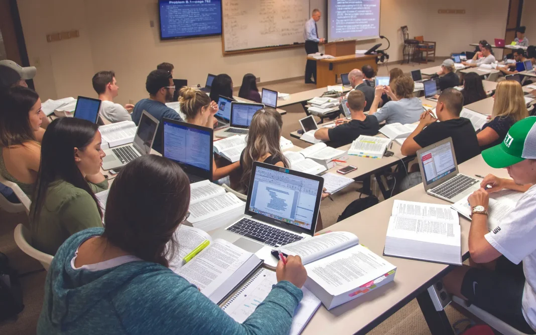 A large university classroom filled with students using laptops and textbooks while a professor lectures at the front near a whiteboard and presentation screens.