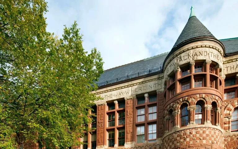 Historic red-brick Harvard Law School building with arched windows and a turreted corner tower, partially framed by leafy trees.