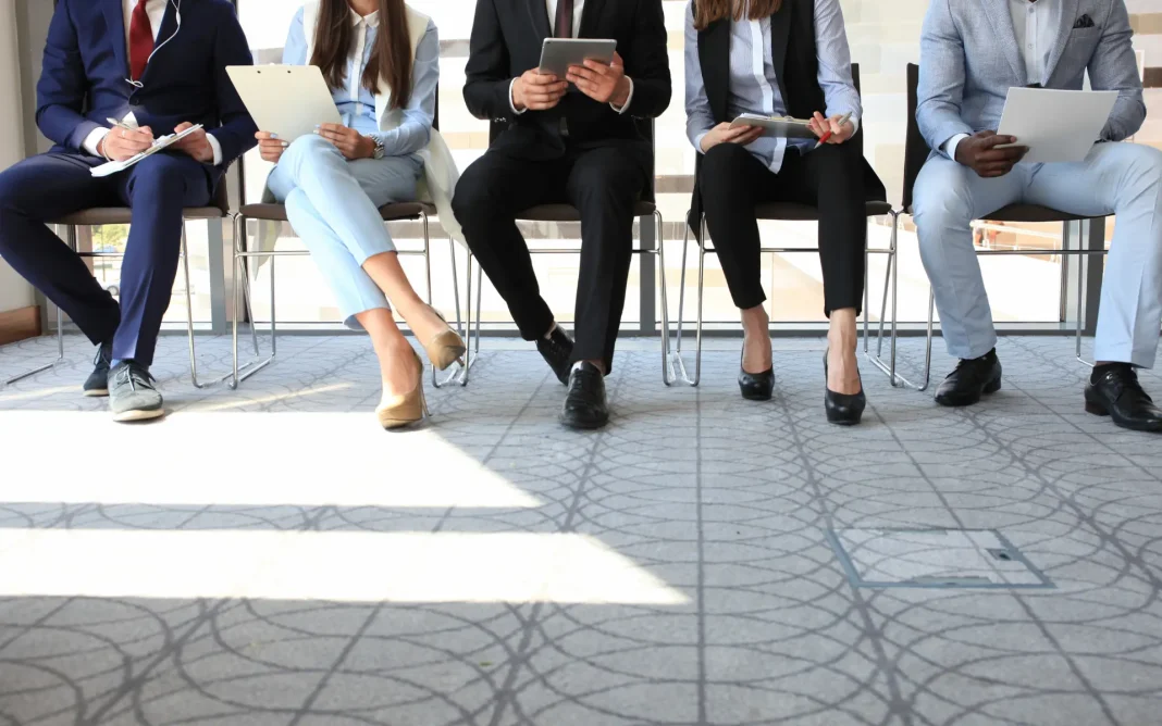 Five professionally dressed people seated in a row holding clipboards, tablets, and papers, appearing to wait for interviews.
