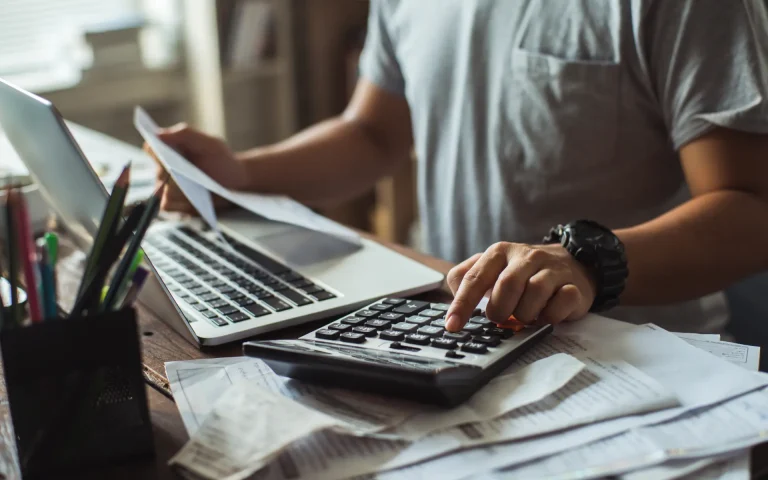 Person reviewing papers and using a calculator beside a laptop on a desk covered with receipts and documents.