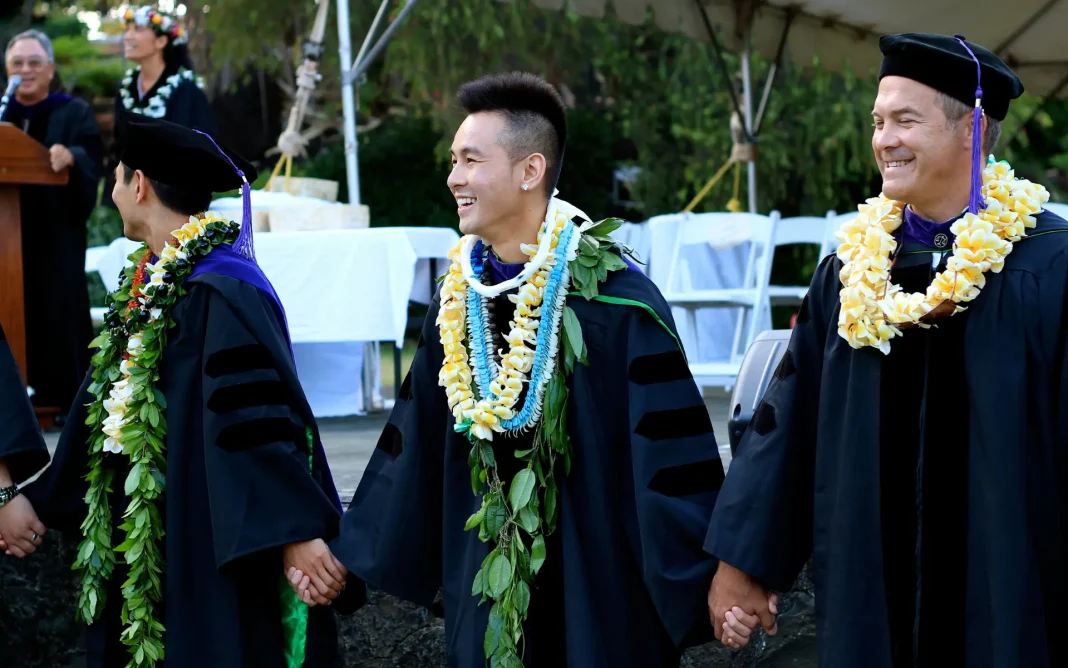 Three graduates in academic gowns and caps wearing flower leis stand hand in hand and smile during an outdoor graduation ceremony.