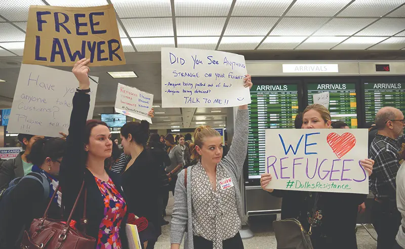 A group of people at an airport arrivals area holding signs offering legal help and support for refugees, including messages like “Free Lawyer” and “We Love Refugees,” indicating advocacy and assistance efforts.