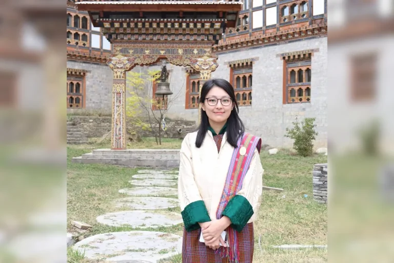 A woman wearing traditional attire and glasses stands in a courtyard in front of an ornate Bhutanese-style building with colorful carved wood details and a hanging bell.