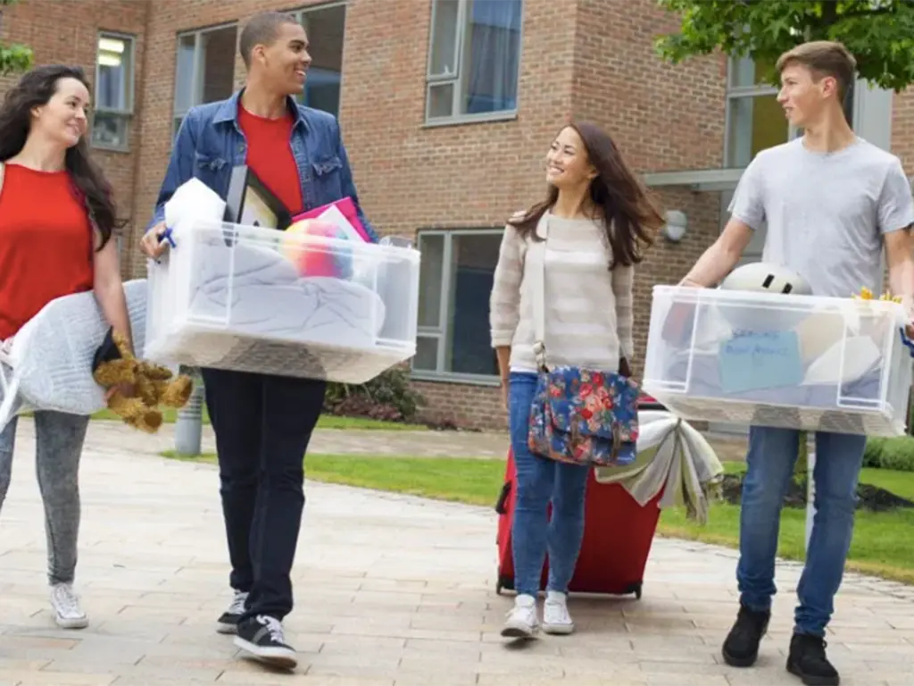 A group of college students walking together outside a campus residence building while carrying clear storage bins and belongings, appearing to move into a dorm at the start of the school year.