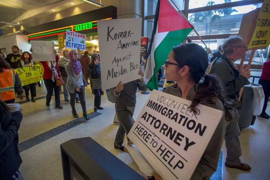 A group of protesters inside a public building holding signs and flags, including messages supporting refugees and opposing a Muslim ban, with one person holding a sign offering help from an immigration attorney.