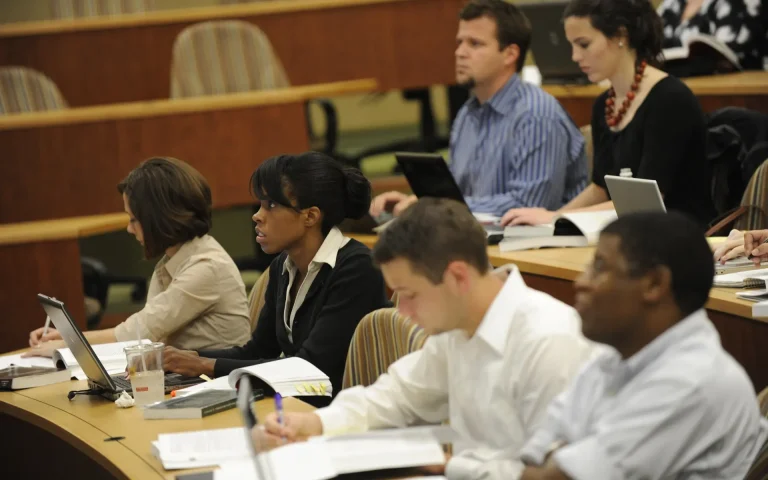 Diverse group of graduate students seated in a tiered lecture hall classroom, taking notes and using laptops during a law school lecture.