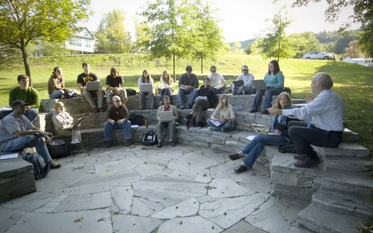 College students seated in a semicircle outdoor classroom while a professor leads a discussion, with many students using laptops and notebooks in a campus setting.